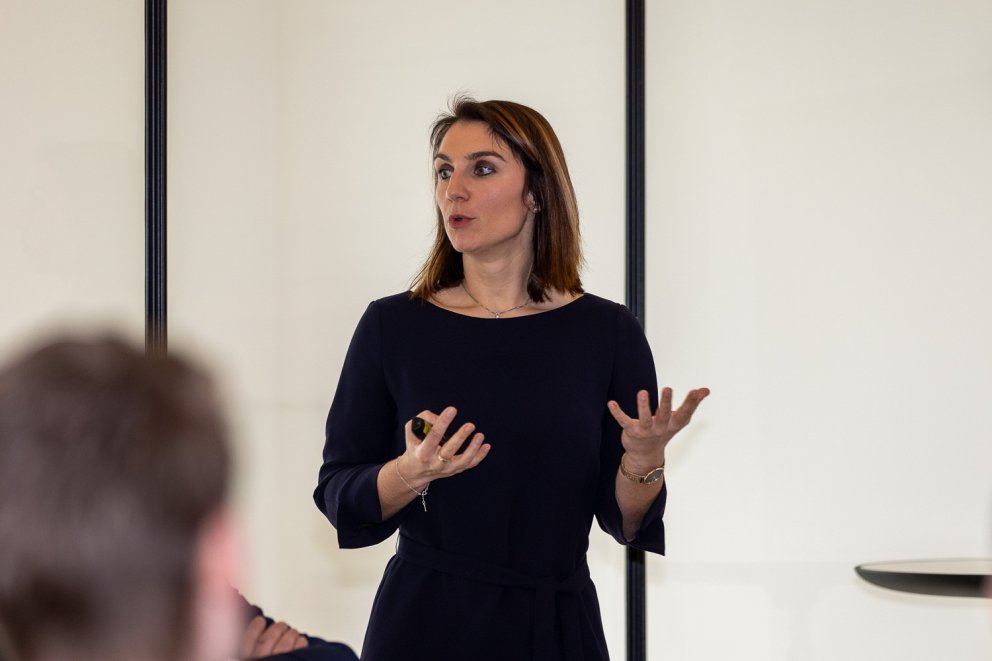 Vrouw in een zwarte jurk die aan het presenteren is, met haar handen uitgestrekt en een gezicht dat aandachtig spreekt.